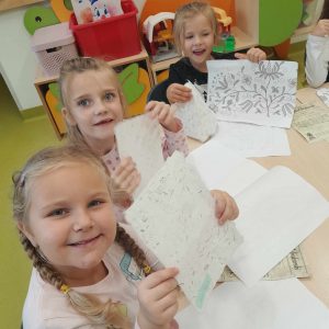 Trzy uczennice oddziału przedszkolnego siedzą przy wspólnym stole. Dziewczynki się uśmiechają i trzymają przed sobą kartki z czarnymi nadrukami. na stole widać rozłożone kartki papieru.
