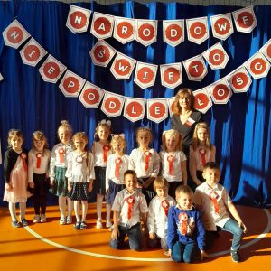 Uczniowie oddziału przedszkolnego pozują do wspólnego zdjęcia ze swoją wychowawczynią. Dzieci mają przypięte do bluzek kotyliony. Za nimi na niebieskim tle widnieje napis: "NARODOWE ŚWIĘTO NIEPODLEGŁOŚCI".