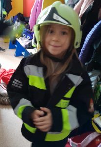 Uczennica oddziału przedszkolnego pozuje do zdjęcia w strażackiej kurtce i hełmie na głowie.