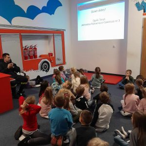 Uczniowie oddziału przedszkolnego siedzą na podłodze. Dzieci patrzą w stronę ściany na której wyświetlany jest slajd z napisem : "Dzień dobry ! Ognik Toruń Jednostka Ratowniczo-Gaśnicza nr 3".