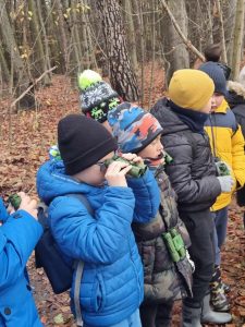 Uczniowie stoją w lesie. Mają przy sobie zielone lornetki. Jeden z chłopców przykłada swoją do oczu.