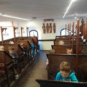 Uczniowie siedzą za ustawionymi w dwóch rzędach stołami z podniesionymi pod skosem blatami.