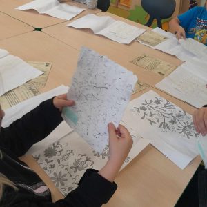 Zdjęcie stołu na którym rozłożone są zadrukowane kartki-pamiątki z muzeum druku i piśmiennictwa. Na pierwszym planie widać dłonie uczennicy w których trzyma kartkę ze znakami po druku.