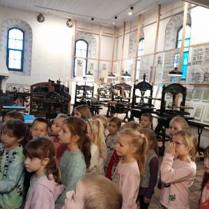 Uczniowie stoją w pomieszczeniu muzeum. Za nimi widać różnego rodzaju prasy drukarskie i ściany ozdobione gotowymi nadrukami.