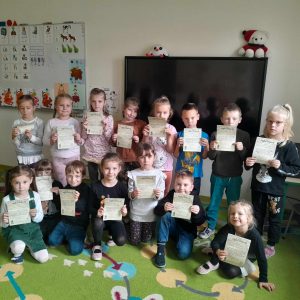 Uczniowie oddziału przedszkolnego pozują do wspólnego zdjęcia na dywanie. Dzieci trzymają przed sobą otrzymane w muzeum piśmiennictwa dyplomy.