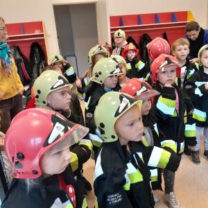 Uczniowie oddziału przedszkolnego pozują do zdjęcia w strażackich kurtkach. Dzieci na głowie mają jasnozielone i czerwone hełmy.