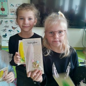 Dwie uczennice stoją na dywanie. Dziewczynki trzymają w dłoniach trzymają swoje pamiątki z muzeum druku i piśmiennictwa.