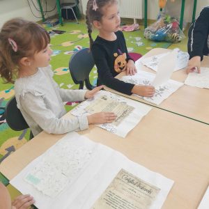 Dwie uczennice oddziału przedszkolnego siedzą przy wspólnym stole. Przed nimi leżą kartki papieru z czarnymi nadrukami.