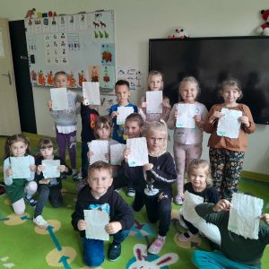 Uczniowie oddziału przedszkolnego pozują do wspólnego zdjęcia na dywanie w swojej sali. Dzieci trzymają przed sobą kartki z nierównymi brzegami.