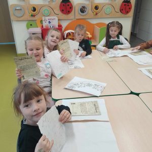 Uczniowie oddziału przedszkolnego siedzą przy wspólnym stole. Dzieci podnoszą w górę kartki papieru - pamiątki z muzeum piśmiennictwa. Przed nimi na stole leżą kartki z różnymi nadrukami.