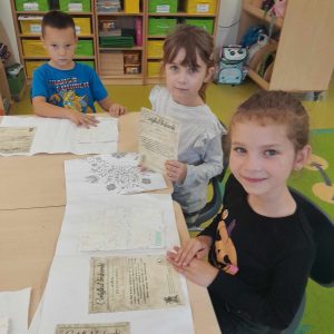 Troje uczniów oddziału przedszkolnego siedzi przy wspólnym stole, na którym rozłożone są pamiątki z wizyty w muzeum druku i piśmiennictwa. Dzieci patrzą w stronę obiektywu.