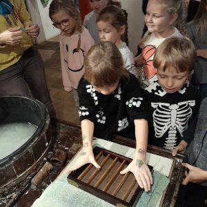 Uczennica opiera dłonie o drewniana skrzyneczkę z równo ułożonymi w sposób równoległy patykami. Obok stoi przyglądający się jej pracy inny uczeń. W tle widać dziewczynki również spoglądające w jej stronę.