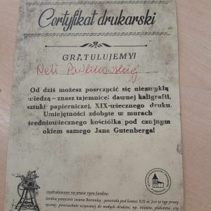 Zdjęcie certyfikatu otrzymanego przez jedną z uczennic. Na jasnym papierze lekko zabrudzonym na czarno napisy: "Certyfikat drukarski", "GRATULUJEMY Neli Pawlikowskiej", "Od dziś możesz poszczycić się niezwykła wiedzą - znasz tajemnice: dawnej kaligrafii, sztuki papierniczej, XIX-wiecznego druku. Umiejętności zdobyte w murach średniowiecznego kościoła pod czujnym okiem samego Jana Gutenberga!". . Poniżej logo muzeum oraz informacja: "Wydrukowano na prawie typu Jardine: Jardine potocznie zwana Bostonką - powstała pod koniec XIX w. Jest to typ prasy ręcznej, powszechnie używanej do małych druków, np. biletów, plakatów czy" w tym miejscu zdjęcie się urywa.