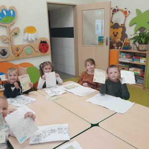 Uczniowie oddziału przedszkolnego siedzą przy wspólnym stole. Dzieci podnoszą w górę kartki papieru - pamiątki z muzeum piśmiennictwa. Przed nimi na stole leżą kartki z różnymi nadrukami.