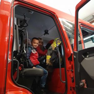 Uczeń oddziału przedszkolnego siedzi w kabinie wozu strażackiego z otwartymi drzwiami przez które widać chłopca. Chłopiec się uśmiecha.