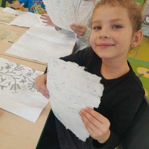 Uczennica oddziału przedszkolnego siedzi przy stoliku. W dłoniach przed sobą trzyma nierówna kartkę z lekkim nadrukiem. Na stole przed nią leżą inne pamiątki z muzeum piśmiennictwa.