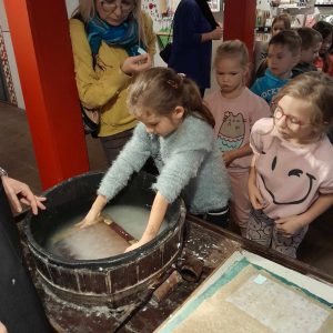 Uczennica zanurza w drewnianej bali z mlecznym płynem deskę. Za nią stoją inni uczniowie przyglądający się tej czynności.