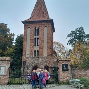 Uczniowie oddziału przedszkolnego stoją przed starym ceglanym budynkiem ze spiczastym dachem. W budynku mieści się muzeum piśmiennictwa.