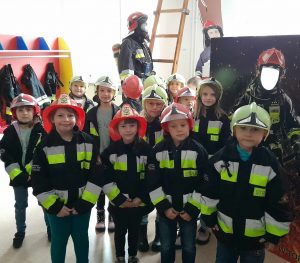 Uczniowie oddziału przedszkolnego pozują do zdjęcia w strażackich kurtkach. Dzieci na głowie mają jasnozielone i czerwone hełmy. W tle widać fototapety ze strażakami i ściankę do pozowania z wyciętym otworem na głowę.