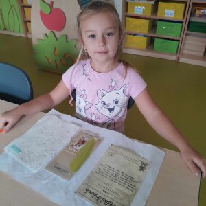 Uczennica oddziału przedszkolnego siedzi przy stoliku na którym opiera dłonie. Przed nią leżą rozłożone pamiątki z muzeum druku i piśmiennictwa.