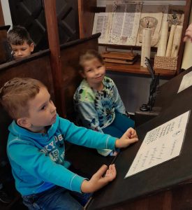 Dwoje uczniów siedzi w jednej ławie przed zamontowanym pod skosem blatem. Chłopiec opiera dłonie o blat, na którym leżą kartki papieru z wykaligrafowanymi literami.