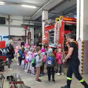 Dzieci z oddziału przedszkolnego stoją przy wozie strażackim. Na pierwszym planie widoczny jest przechodzący strażak.