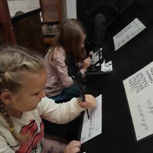 Dwie uczennice pracują przy zamocowanym pod skosem blatem. Dziewczynki trzymają w dłoniach pióra, którymi piszą po leżących przed nimi kartkach. Na blacie ułożone są również kartki z wykaligrafowanymi w różnych czcionkach literami.