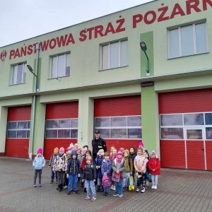 Uczniowie oddziału przedszkolnego pozują do wspólnego zdjęcia z Panem strażakiem przed budynkiem z napisem "PAŃSTWOWA STRAŻ POŻARNA".