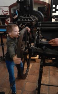 Uczeń kręci dużym metalowym kołem. Chłopiec obejmuje koło szeroko rozstawionymi rękoma. Dla równowagi ma również szeroko rozstawione nogi.