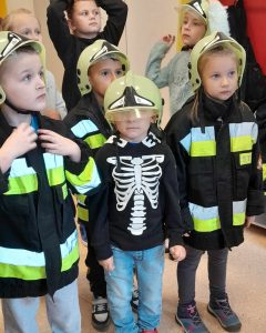 Uczniowie oddziału przedszkolnego pozują do zdjęcia w strażackich kurtkach. Dzieci na głowie maja jasnozielone hełmy.