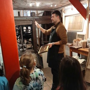 Uczniowie przygladają sie Panu ubranemu w czarny fartuch. Mężczyzna w jednej dłoni trzyma drewnianą deskę, a drugą rękę podnosi w górę.