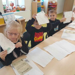 Uczniowie oddziału przedszkolnego siedzą przy wspólnym stole. Dzieci podnoszą w górę kartki papieru - pamiątki z muzeum piśmiennictwa. Przed nimi na stole leżą kartki z różnymi nadrukami.