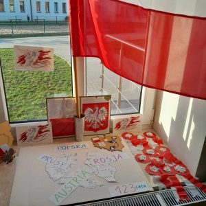 Zdjęcie dekoracji okna sali. W centralnym puncie na parapecie umieszczona jest flaga a pod nią godło Polski, kotyliony oraz tablica z napisami "PRUSY", "ROSJA", "AUSTRIA" "123 LATA" oraz przekreślonym napisem "POLSKA".