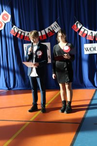 Dwoje uczniów prowadzących apel stoi obok siebie trzymając w dłoniach sztywne podkładki. Chłopiec trzyma w drugiej dłoni mikrofon.