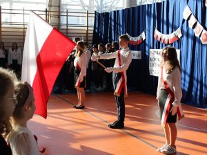 Zdjęcie stojącego na scenie pocztu flagowego szkoły. Dwie dziewczynki i chłopiec stoją w równych odstępach od siebie. Uczniowie maja przewieszone przez ramię biało czerwone szarfy. Chłopiec stojący po środku trzyma opuszczoną w przód flagę Polski.