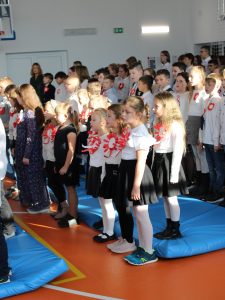 Uczniowie biorący udział w apelu podczas śpiewania hymnu Polski. Dzieci stoją na baczność. Do bluzek mają przyczepione kotyliony.