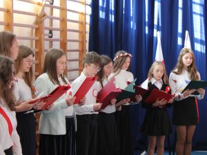 Uczniowie klasy szóstej stoją w jednym rzędzie tworząc chórek. Dzieci trzymają przed sobą sztywne podkładki z tekstem pieśni.