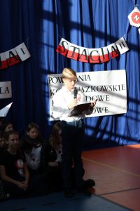 Uczeń klasy szóstej stoi na scenie trzymając w dłoniach mikrofon i sztywną podkładkę z tekstem. W tle widać fragment dekoracji.