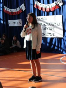 Uczennica klasy szóstej stoi na scenie trzymając przed sobą mikrofon. W tle widać fragment dekoracji.