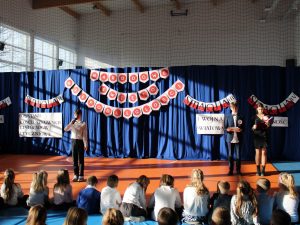 Zdjęcie sceny przygotowanej z okazji apelu. Na pierwszym planie widać siedzących na materacach uczniów. Przed nimi stoi chłopiec z mikrofonem a po jego prawej stronie dwoje uczniów prowadzących apel. Za nimi na niebieskiej kurtynie po środku umieszczony jest napis: "NARODOWE ŚWIĘTO NIEPODLEGŁOŚCI" a po obu jego stronach zamieszczone są napisy "STACJA II" i pod nim "POWSTANIE KOŚCIUSZKOWSKIE LISTOPADOWE I STYCZNIOWE" dalej "STACJA III" a pod nim "I WOJNA ŚWIATOWA" oraz "STACJA III" i pod nim napis "WOLNOŚĆ".