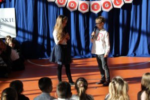 Dwoje uczniów klasy drugiej stoi na scenie na przeciwko siebie. Dzieci w dłoniach trzymają mikrofony.