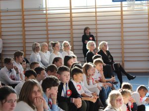 Zdjęcie uczniów siedzących na widowni podczas apelu. W tle widać również siedzących nauczycieli oraz Panią Dyrektor i Sekretarz Gminy Łysomice.