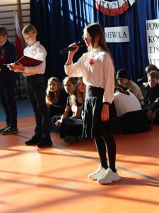 Na scenie stoi uczennica trzymająca przed sobą mikrofon. W tle widać stojącego ucznia trzymającego sztywną podkładkę i siedzących za nim jego kolegów z klasy.