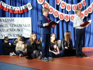 Dwóch uczniów stoi na scenie trzymając przed sobą sztywne podkładki. Za nimi na podłodze siedzą ich koledzy. W tle widać fragment dekoracji.