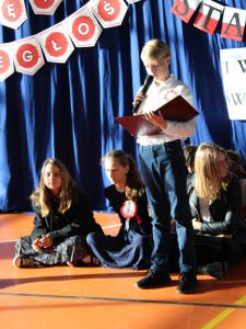 Uczeń stoi z mikrofonem na scenie. Chłopiec trzyma przed sobą sztywną podkładkę. Za nimi widać siedzące na podłodze uczennice.