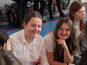 Dwie uczennice klasy ósmej pozują do wspólnego zdjęcia siedząc na ławce śród innych uczniów. Dziewczynki się uśmiechają.