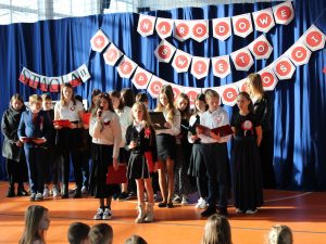 Uczniowie klasy szóstej zgromadzeni podczas występu na sali gimnastycznej. Część z nich trzyma podkładki z tekstem. Dwie dziewczynki stojące po środku trzymają w dłoniach mikrofony. W tle widać zawieszoną na granatowej tkaninie dekorację z napisem "NARODOWE ŚWIĘTO NIEPODLEGŁOŚCI".