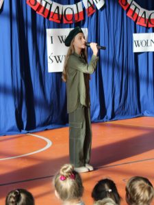 Uczennica w zielonym stroju przypominającym mundur i w zielonym berecie na głowie podczas występu. Dziewczynka trzyma przed sobą mikrofon.