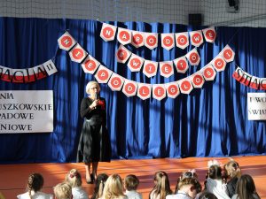 Pani Dyrektor podczas wystąpienia na apelu. Pani trzyma w dłoni mikrofon a przed nią na podłodze siedzą uczniowie. W tle wdać dekoracje - na niebieskiej zasłonie po środku zawieszony napis: "NARODOWE ŚWIĘTO NIEPODLEGŁOŚCI".