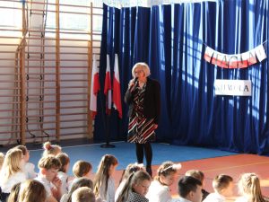 Sekretarz gminy podczas przemówienia na apelu. Pani trzyma w dłoni mikrofon i lekko pochyla się w kierunku siedzących przed nią dzieci.